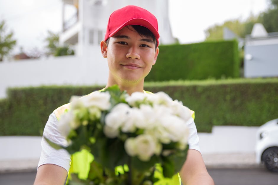 A delivery person wearing a red cap and a light-colored t-shirt holds a bouquet of fresh white flowers, including roses and other blossoms, wrapped in clear cellophane. The flowers exhibit vibrant, he