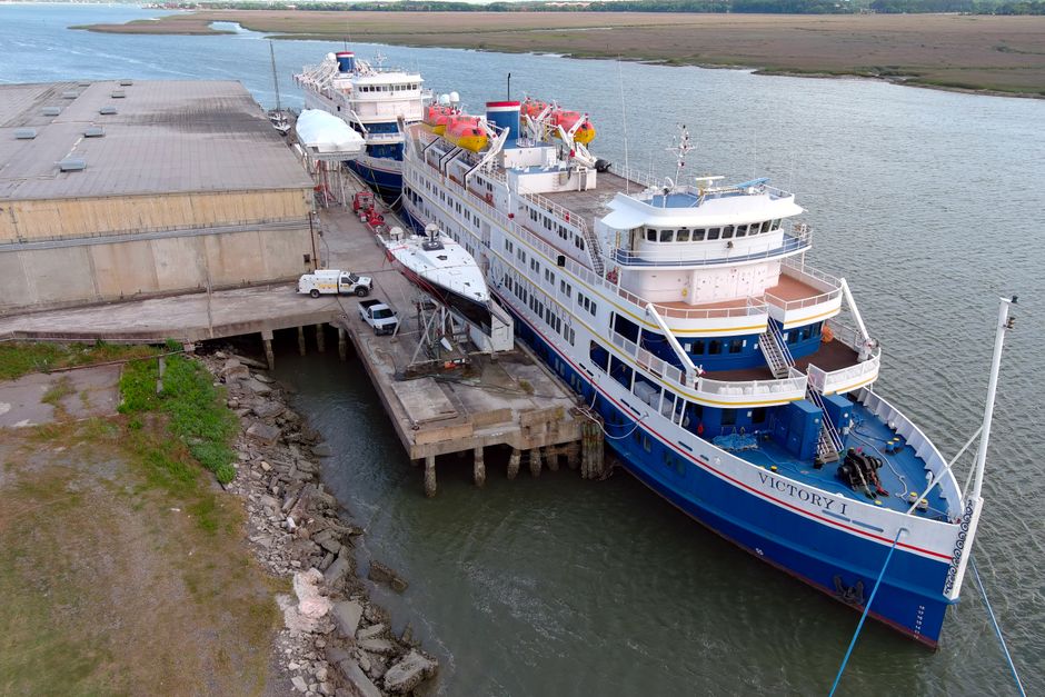 An aerial view of a large white and blue vessel named Victory I docked at a port with surrounding industrial buildings and vehicles, with calm water nearby. The ship has multiple decks with windows an