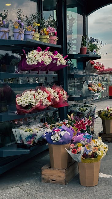 A display of fresh floral arrangements on black metal shelves inside a flower shop with large glass windows. The arrangement includes various bouquets wrapped in colorful paper and ribbons, featuring