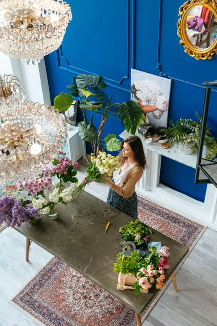 A woman with long brown hair, dressed in a beige top and patterned skirt, is arranging a bouquet of fresh flowers on a dark grey tabletop inside a flower shop with vibrant blue walls. The bouquet incl
