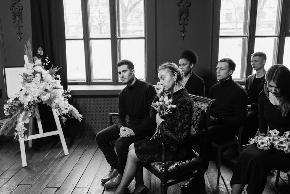 A group of six people seated indoors during a somber occasion, with a large, elegant floral arrangement on an easel to the left featuring white roses, lilies, and greenery, symbolizing sympathy. The w