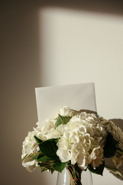 A bouquet of fresh white hydrangeas and roses arranged in a clear glass vase, accented with dark green leaves. The bouquet is partially wrapped with white paper and tied with a ribbon, positioned on a